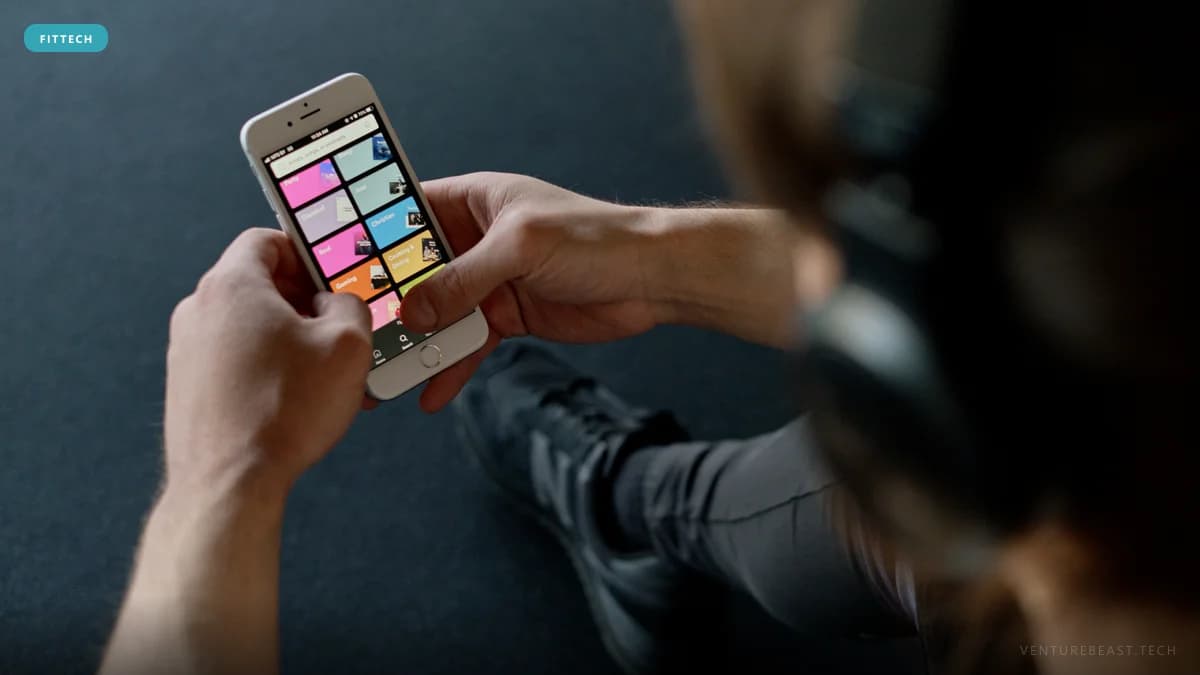 Person in sneakers and headphones sitting on a gym floor, browsing a colorful workout app on a phone
