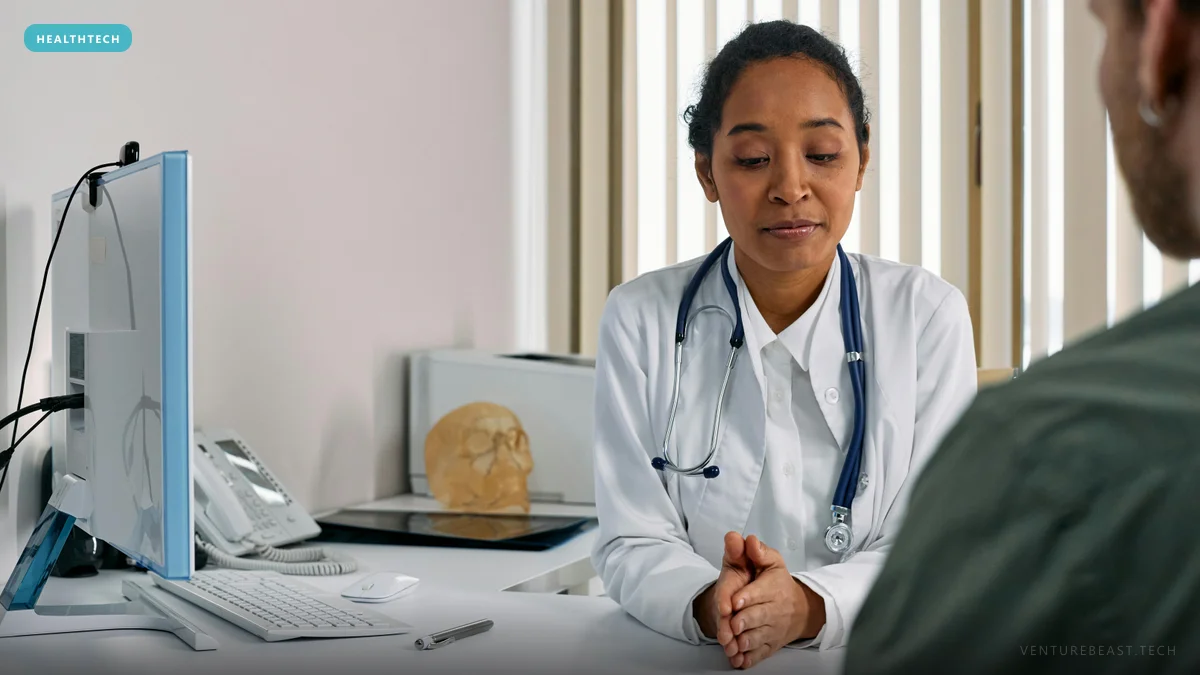 Doctor in a white coat and stethoscope listens to a patient across her desk beside a computer monitor