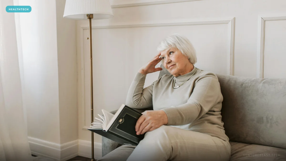 Older woman with white hair sitting on a gray sofa holding a book, resting her head on her hand