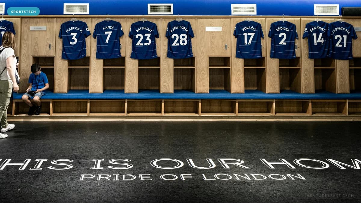 Football dressing room with numbered blue jerseys over lockers and floor text reading "This Is Our Home, Pride of London"