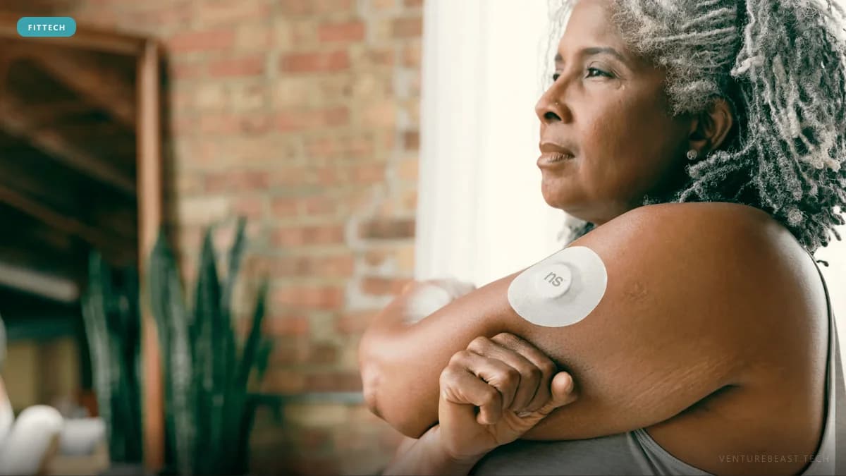 Older woman with gray locs stretching her arm, a round glucose sensor patch on her upper arm