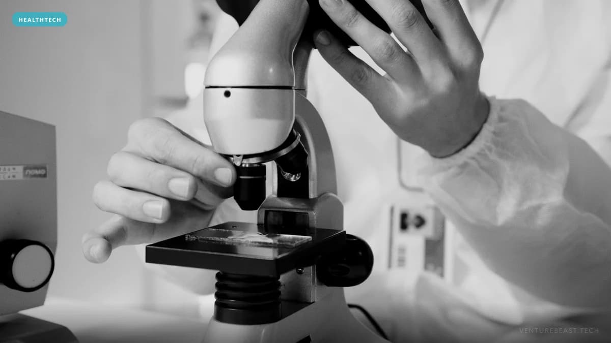 Black-and-white close-up of a lab worker in a protective gown adjusting a microscope over a glass slide