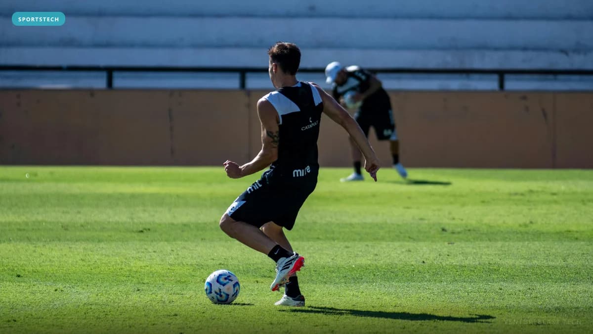 A soccer player in a black training vest dribbling a ball on a sunlit pitch while a teammate warms up behind