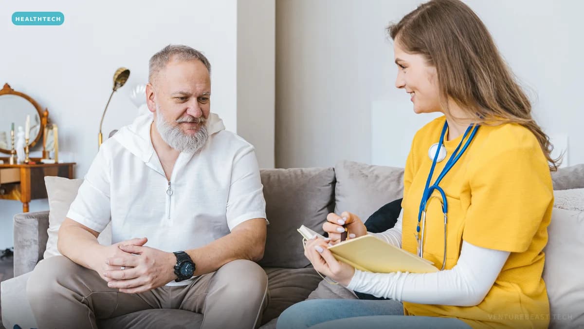 A clinician in yellow scrubs taking notes while talking with an older man on the sofa in his living room