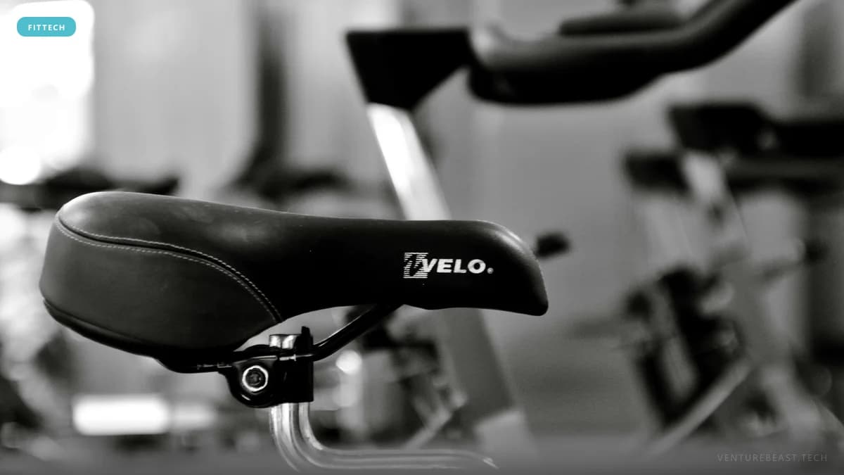 Black-and-white close-up photo of an exercise bike saddle marked VELO, with more indoor bikes blurred behind it