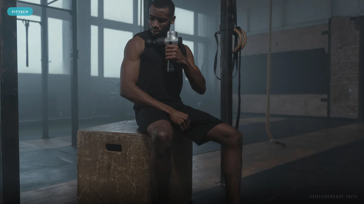 Athlete sitting on a wooden plyo box in a dim gym, using a massage gun on his shoulder