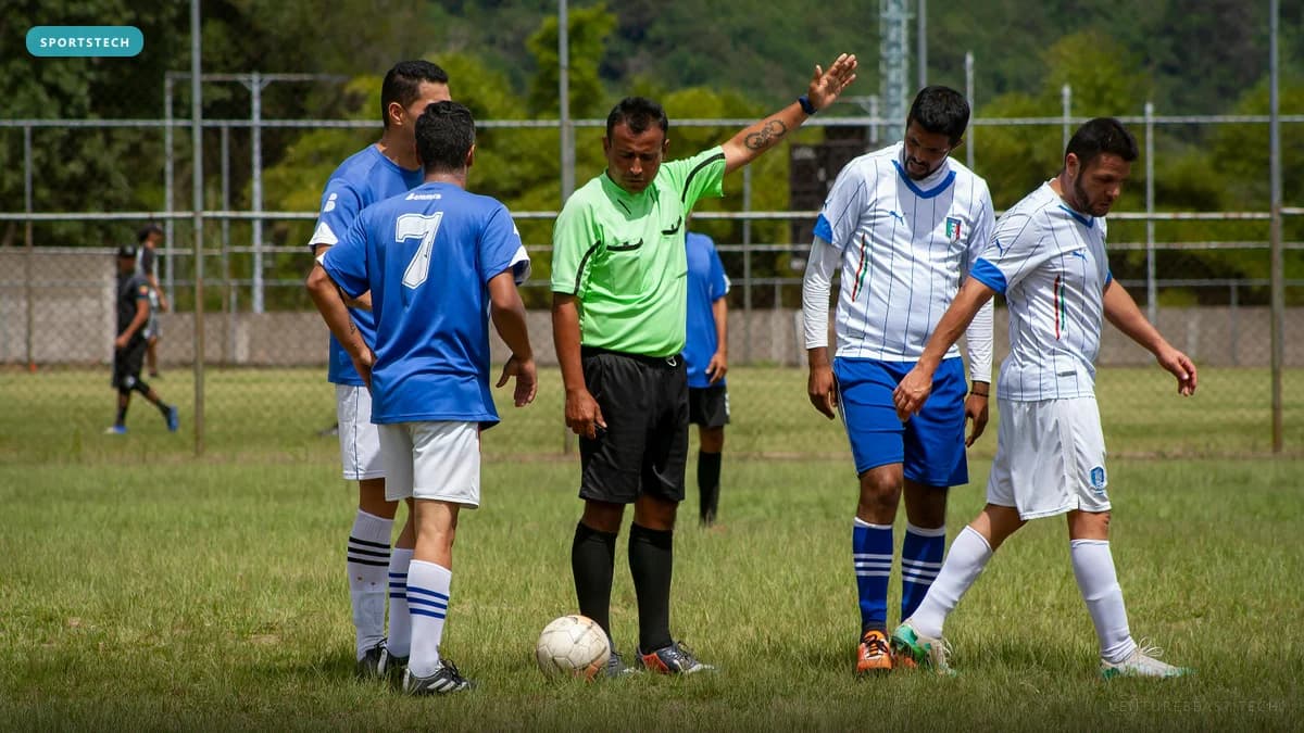 Referee in a green shirt raising his arm between players in blue and white kits on a grass field, ball at his feet