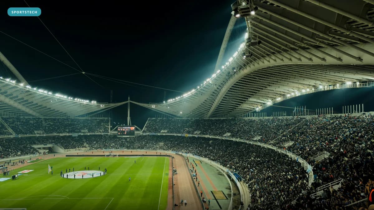 Night view of a packed stadium under a curved, lit roof, overlooking a floodlit soccer pitch and running track