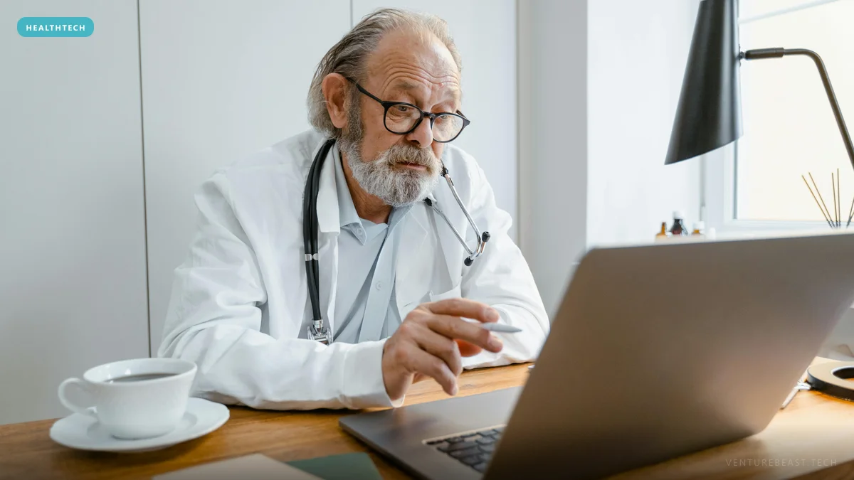 Older bearded doctor in a white coat and stethoscope working at a laptop on a wooden desk beside a cup of coffee